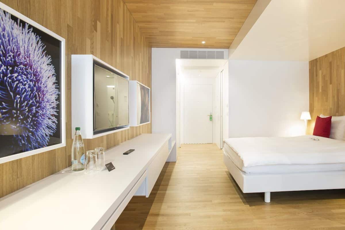 A modern hotel room with a wooden ceiling, a large bed with white bedding, a bedside table with a lamp, and a TV mounted on the wall.