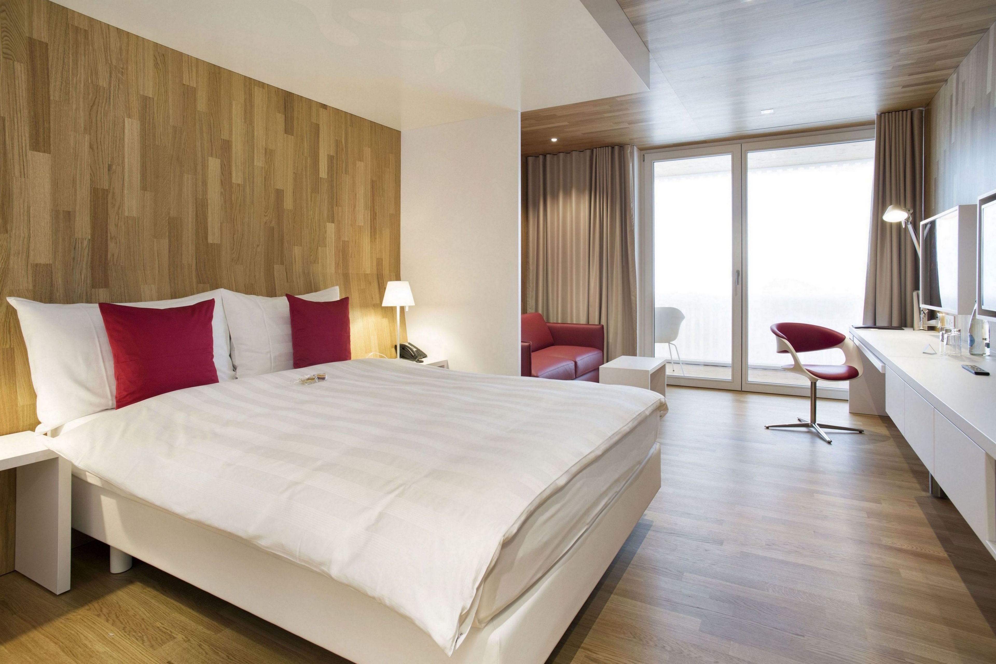 A modern hotel room with a large bed, a desk, a chair, and a balcony with curtains.