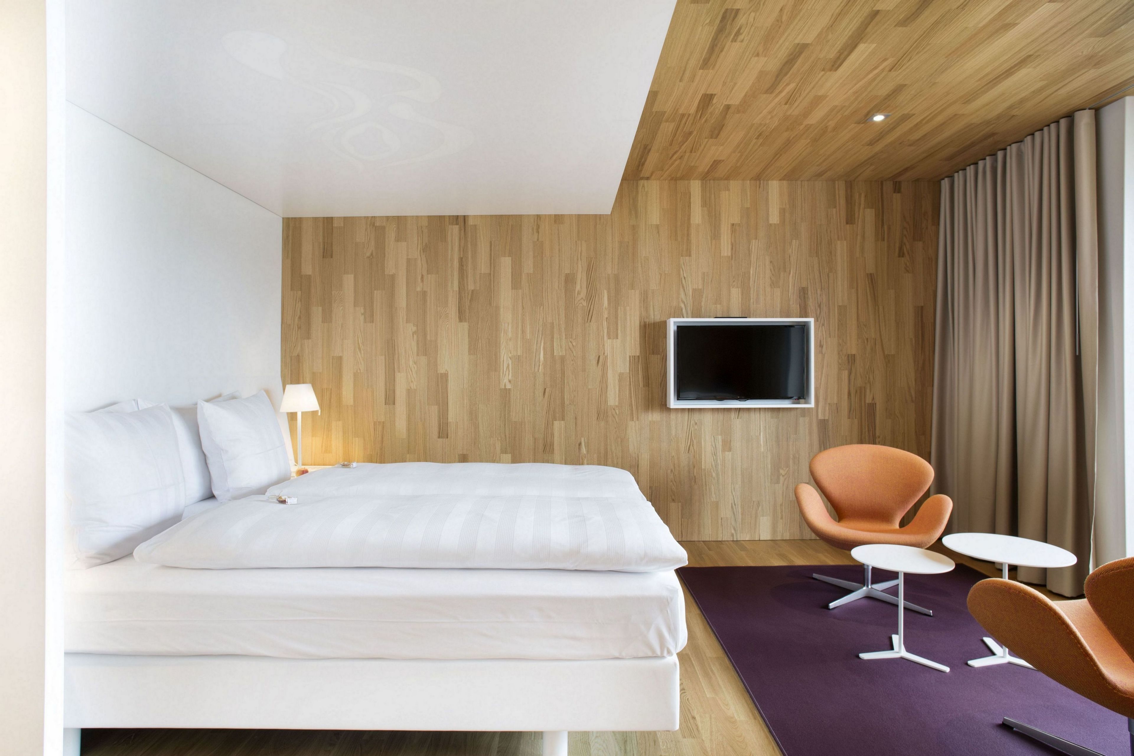 A modern hotel room with a wooden wall, a flat-screen TV, a bed, two orange chairs, and a small table.
