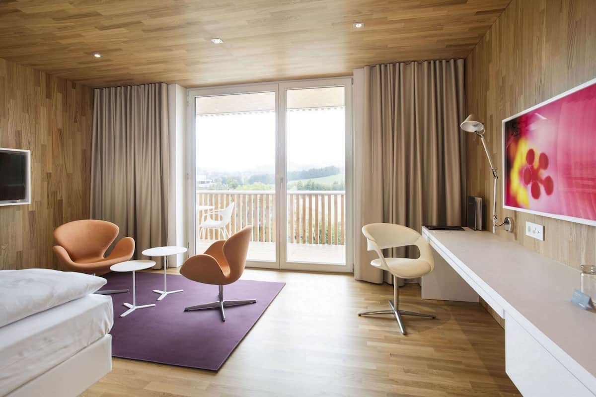 A modern hotel room with a wooden ceiling, a balcony with a view, a sofa, two armchairs, a small table, and a TV.
