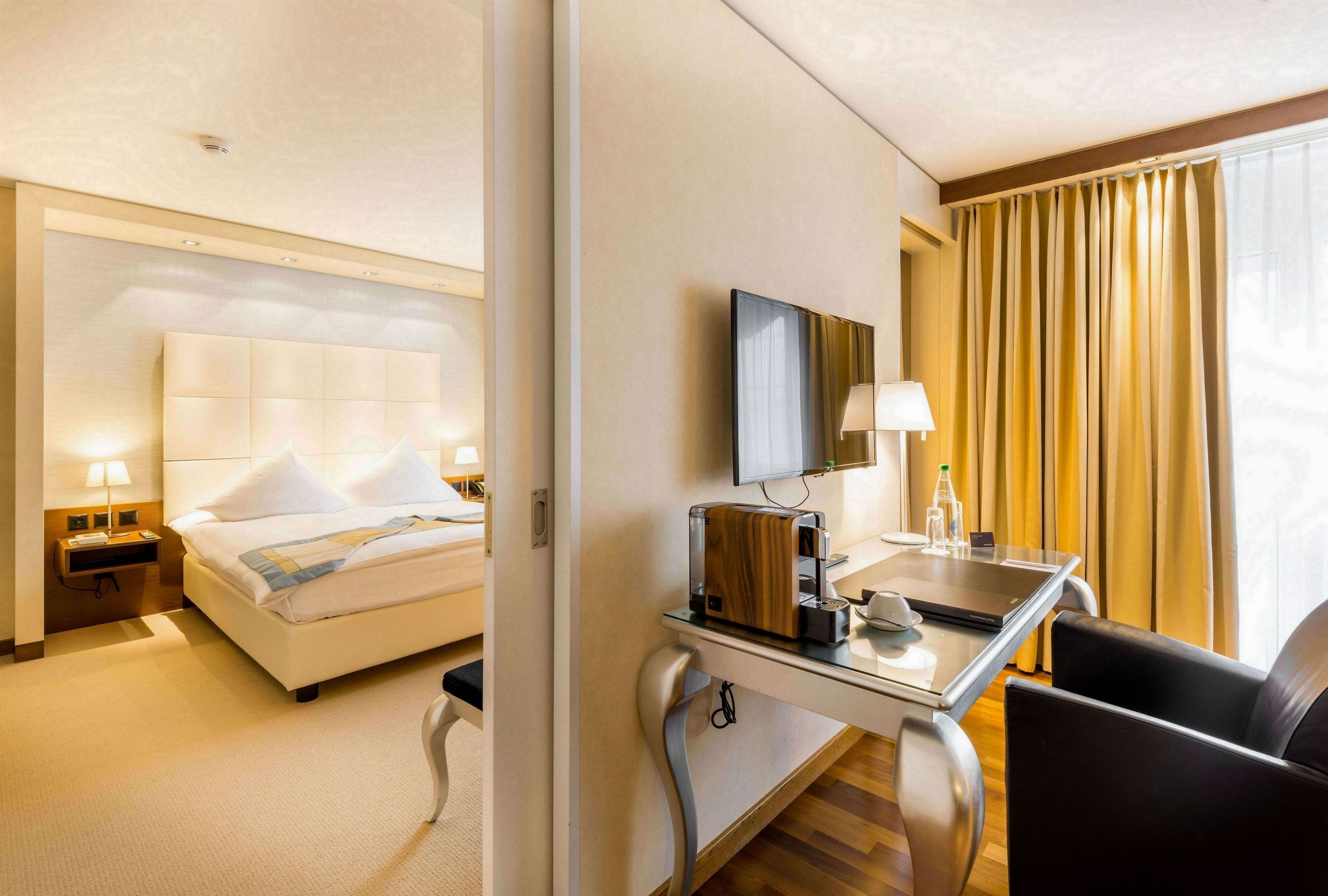 A hotel room with a bed, bedside table with a lamp, a desk with a laptop, and a chair.