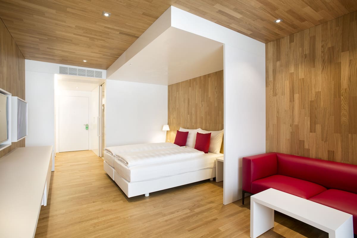 A modern hotel room with a wooden floor, a white bed with red pillows, a red sofa, and a white nightstand.
