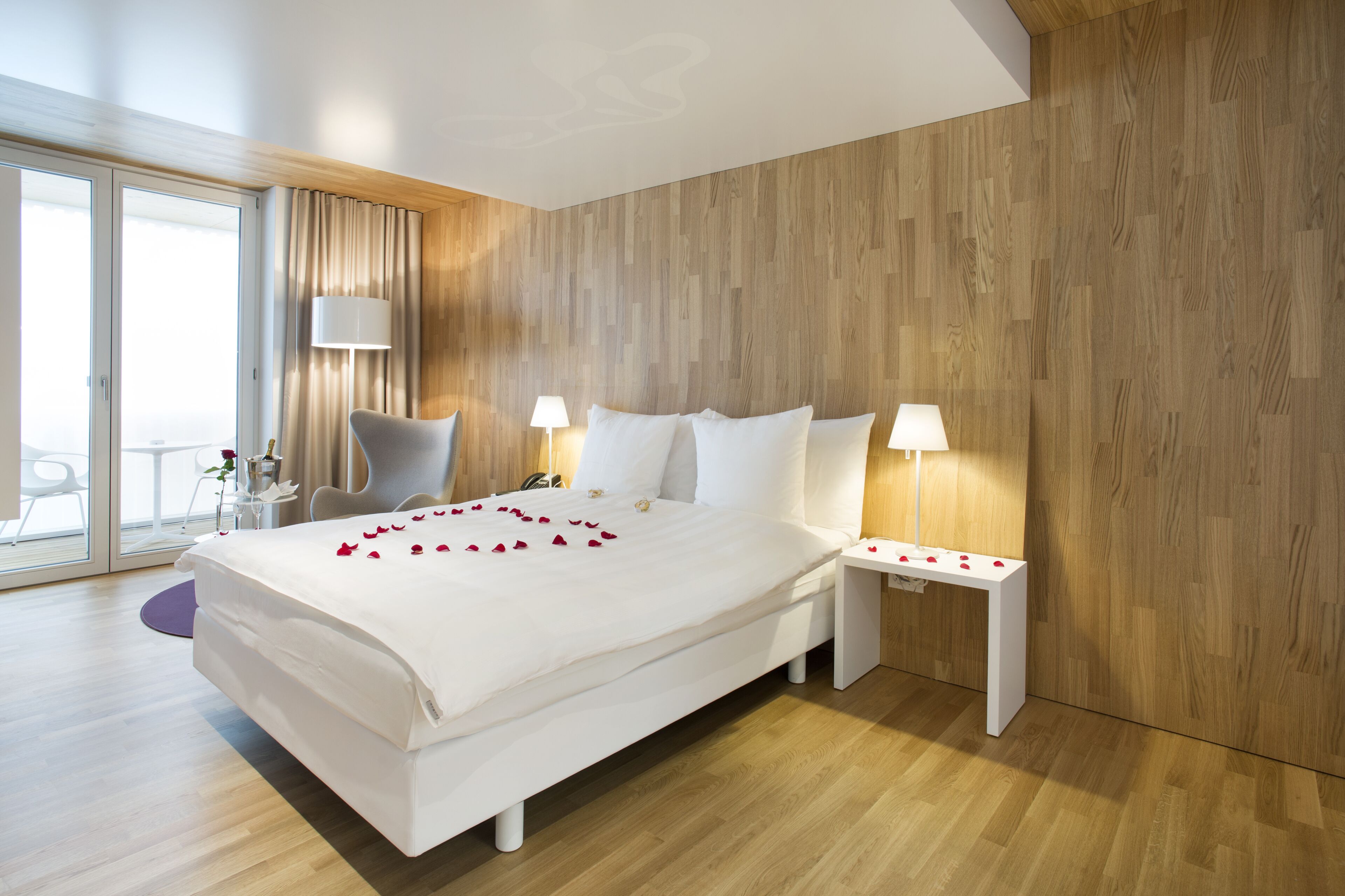 A modern hotel room with a large bed, two bedside tables, a chair, and a view of the outdoors.
