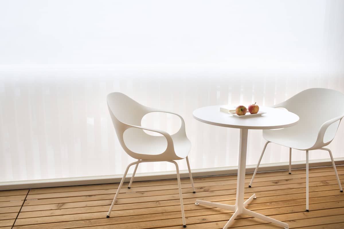 A room with a wooden floor, white chairs, a small round table, and a window with blinds.