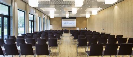Un salón de conferencias con filas de sillas, una pantalla para proyector y una pared de madera.