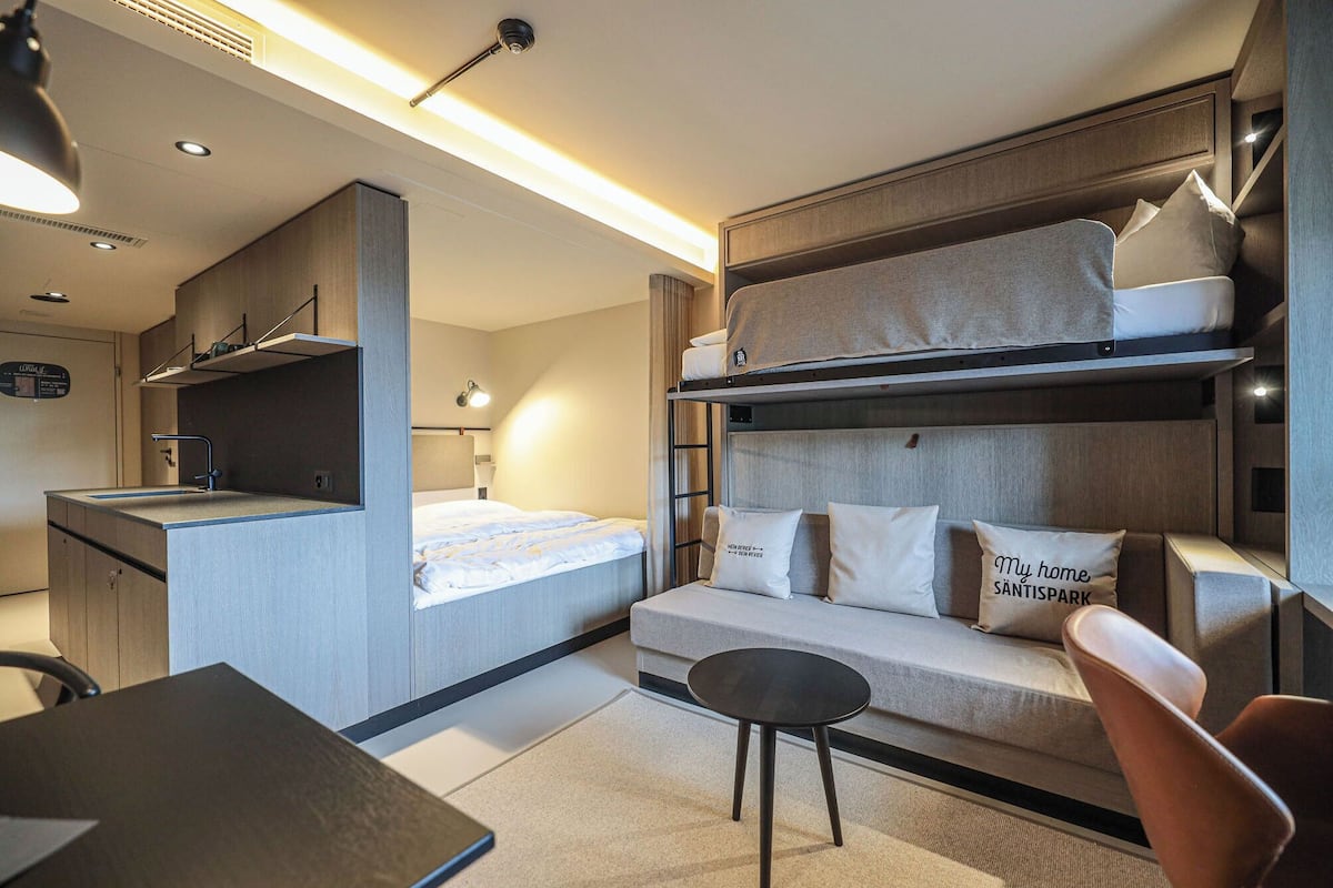 A compact hotel room with bunk beds, a sofa, a small table, and a kitchenette.