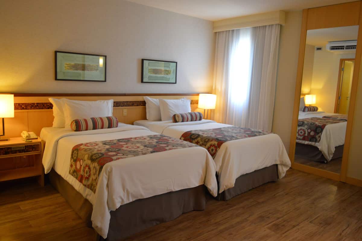 Quarto de hotel com duas camas, uma mesa de cabeceira com abajur e um espelho refletindo o ambiente.