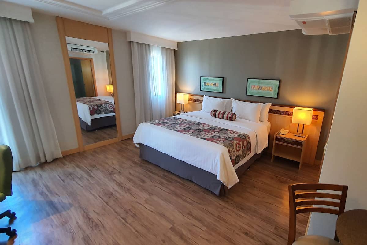 Quarto de hotel com uma cama grande, uma mesa de cabeceira com abajur, uma cadeira e um espelho.