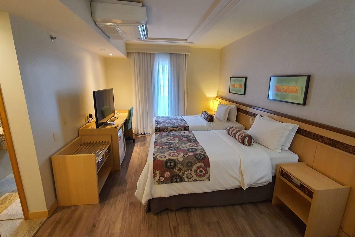 Quarto de hotel com duas camas, uma escrivaninha com computador, televisão e janela com cortinas.