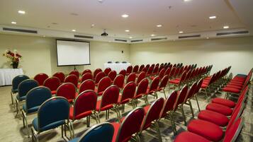 Ein Konferenzraum mit Reihen roter und blauer Stühle, einer Projektionsleinwand und einem Tisch mit weißem Tuch und Blumen.