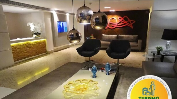 Un moderno vestíbulo de hotel con un mostrador de recepción, dos sillones negros, una mesa de centro de vidrio y una pared decorativa con un diseño abstracto rojo.