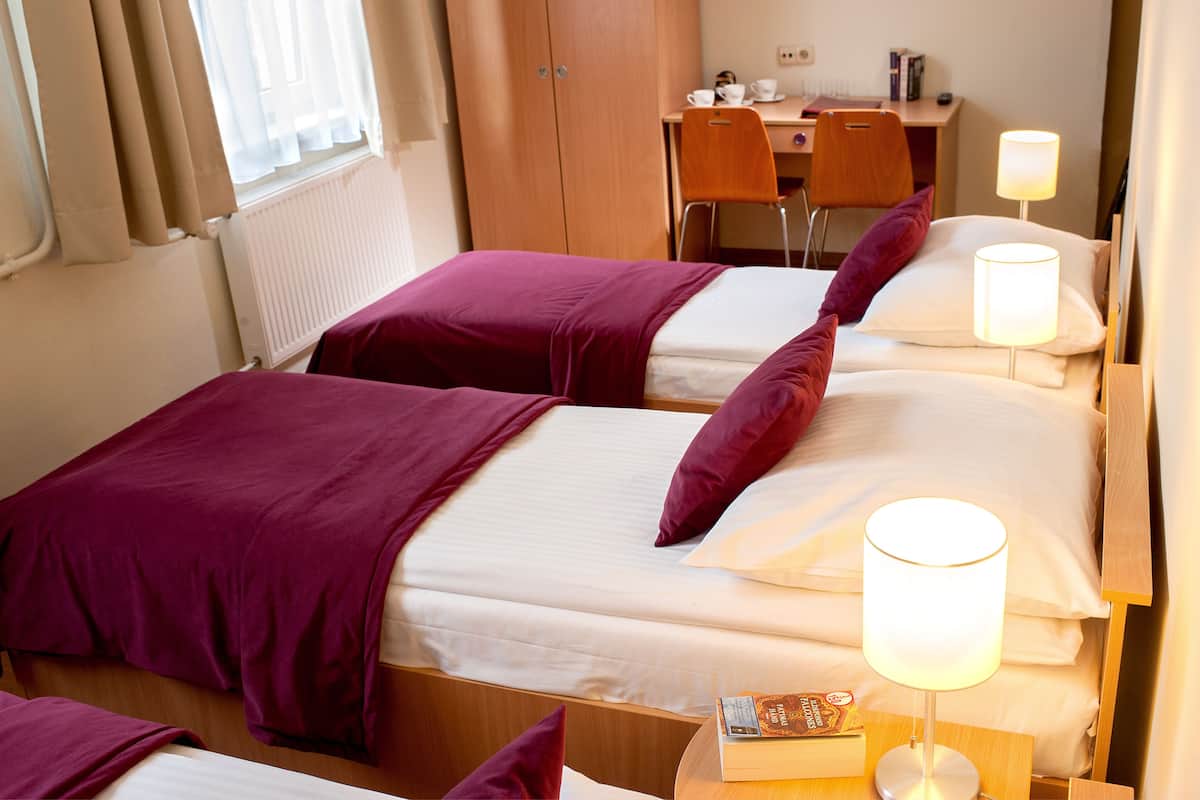 A hotel room with two beds, each with a purple blanket and white pillows, a bedside table with a lamp, and a desk with a chair and a coffee cup.