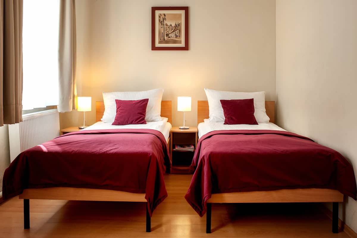 A hotel room with two single beds, each with a wooden frame and red bedding. There are bedside tables with lamps, a framed picture on the wall, and a window with curtains.