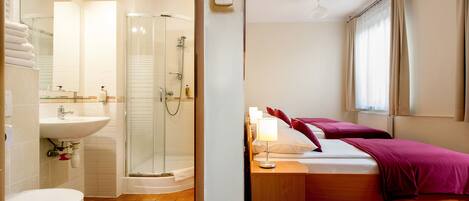 A bathroom with a toilet, sink, and shower. There is a bed with a nightstand and a window with curtains.