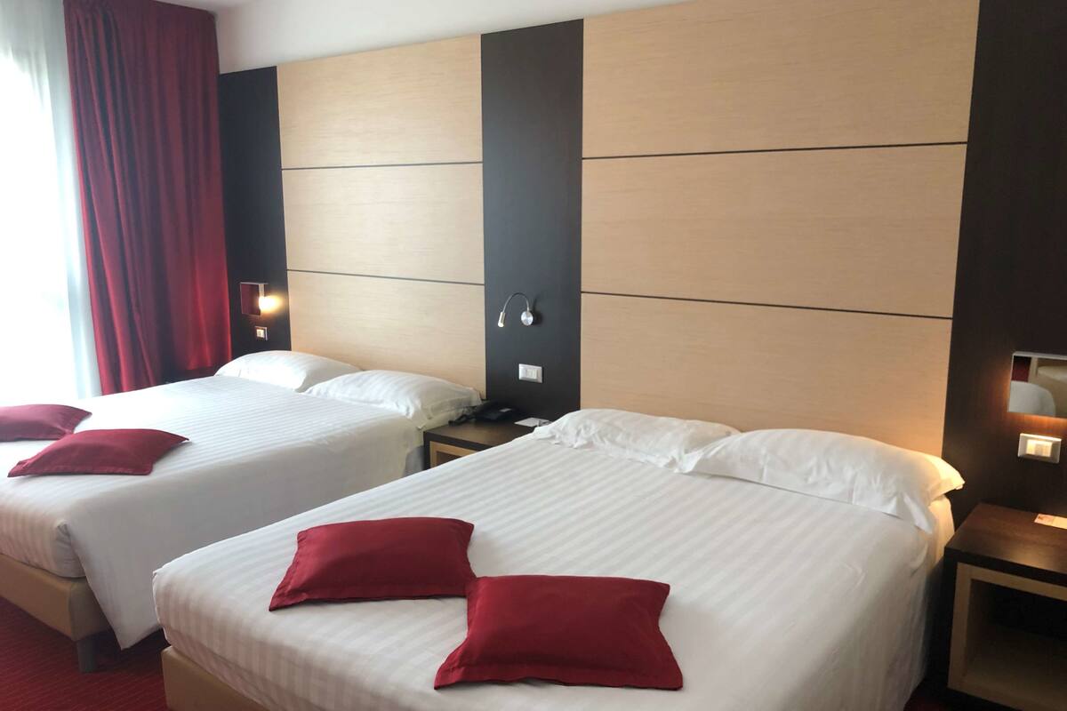 Une chambre d’hôtel avec deux lits, chacun agrémenté de draps blancs et d’oreillers rouges, une tête de lit en bois, des lampes de chevet et un rideau rouge.