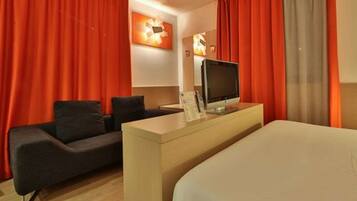 A hotel room with a bed, a desk with a flat-screen TV, a sofa, and red curtains.