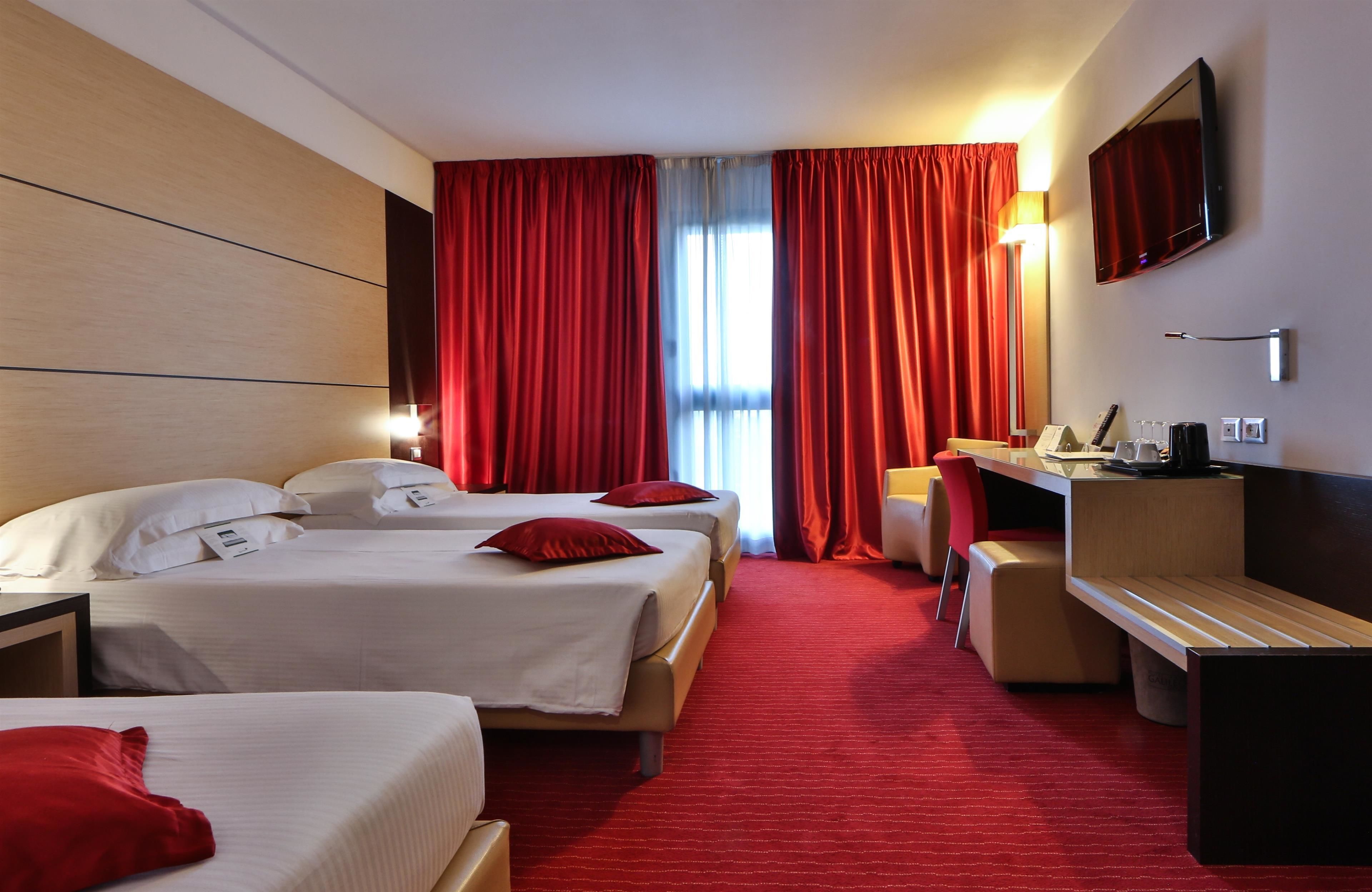 A hotel room with two beds, red curtains, a desk, a chair, and a TV mounted on the wall.