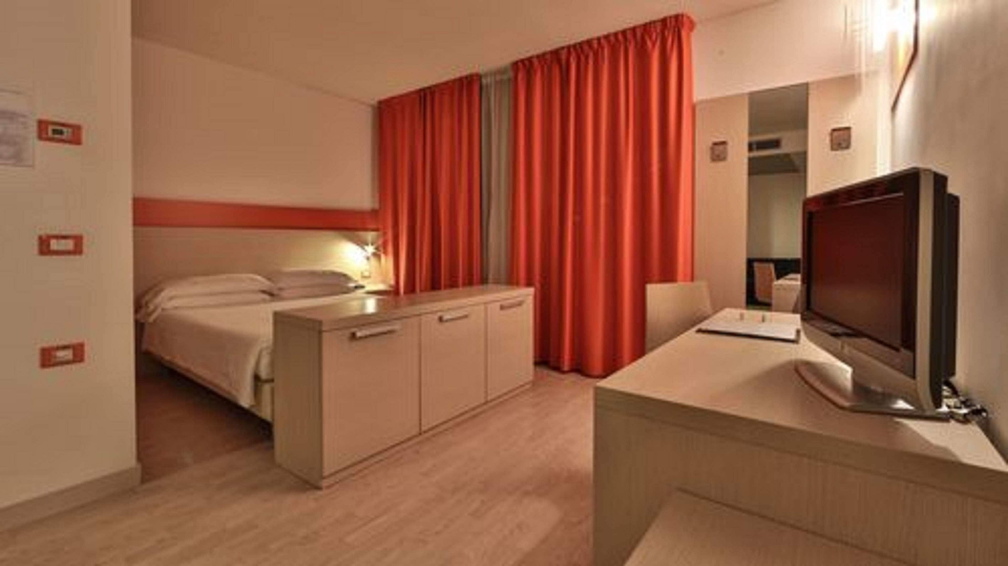 A hotel room with a bed, a desk with a computer, a TV, and a window with red curtains.