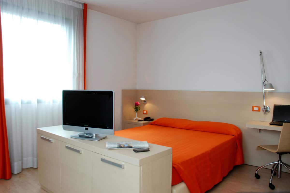 Une chambre d’hôtel avec un lit, un bureau avec un ordinateur, une lampe et une fenêtre avec des rideaux.