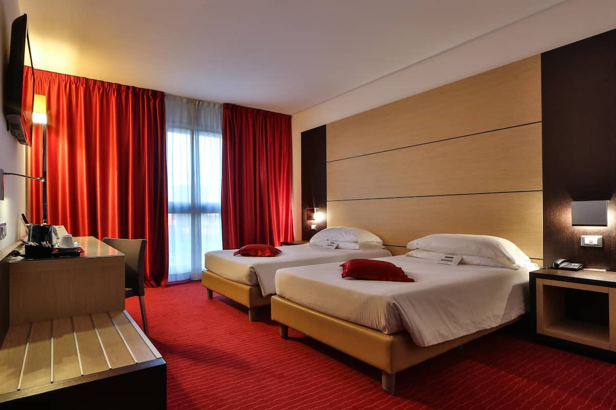 Une chambre d’hôtel avec deux lits, un bureau avec une chaise, une petite table et une moquette rouge.