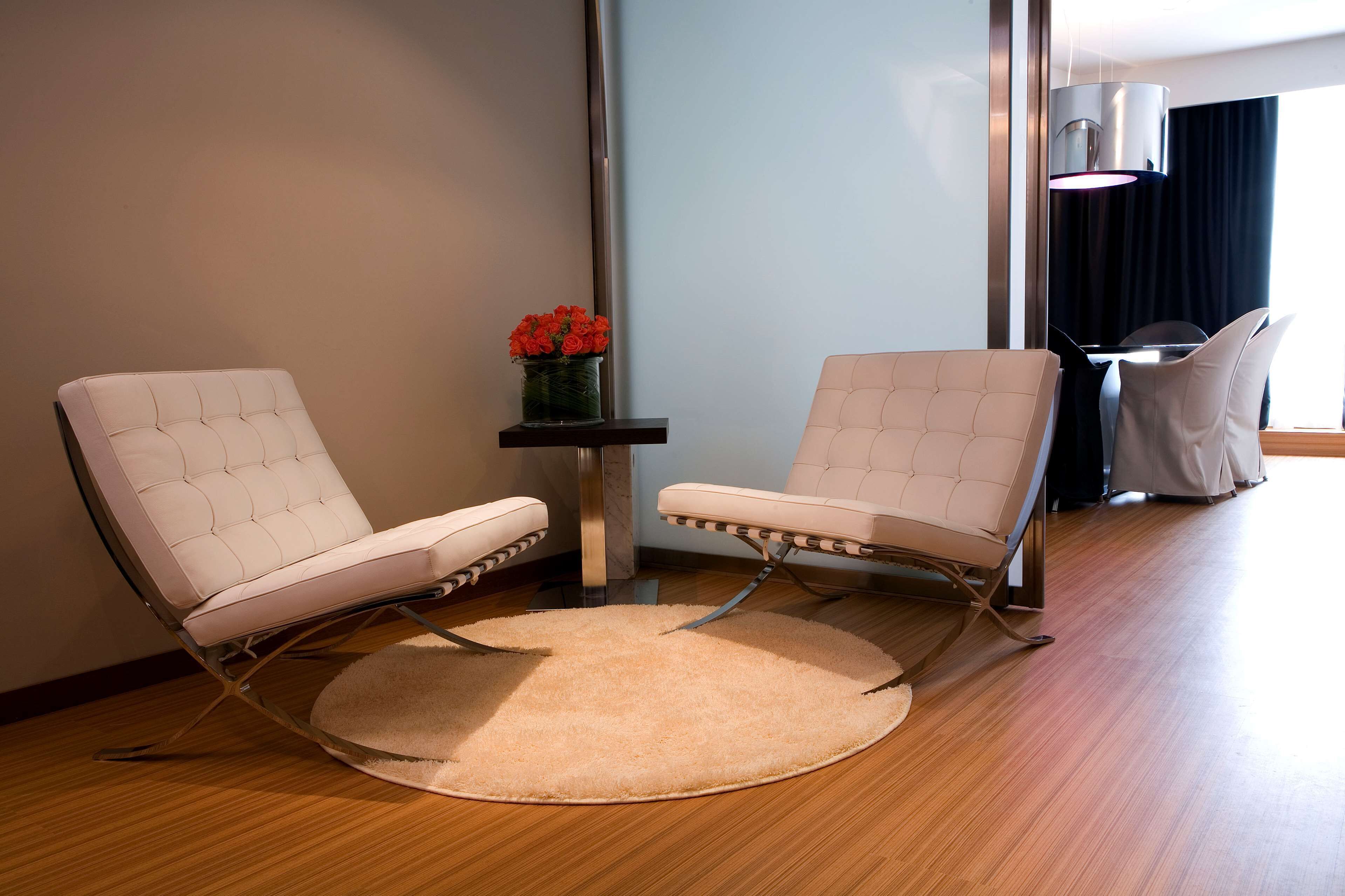 Una habitación moderna con dos sillones de cuero blanco, una mesita con un jarrón de flores rojas y una alfombra redonda sobre un suelo de madera.