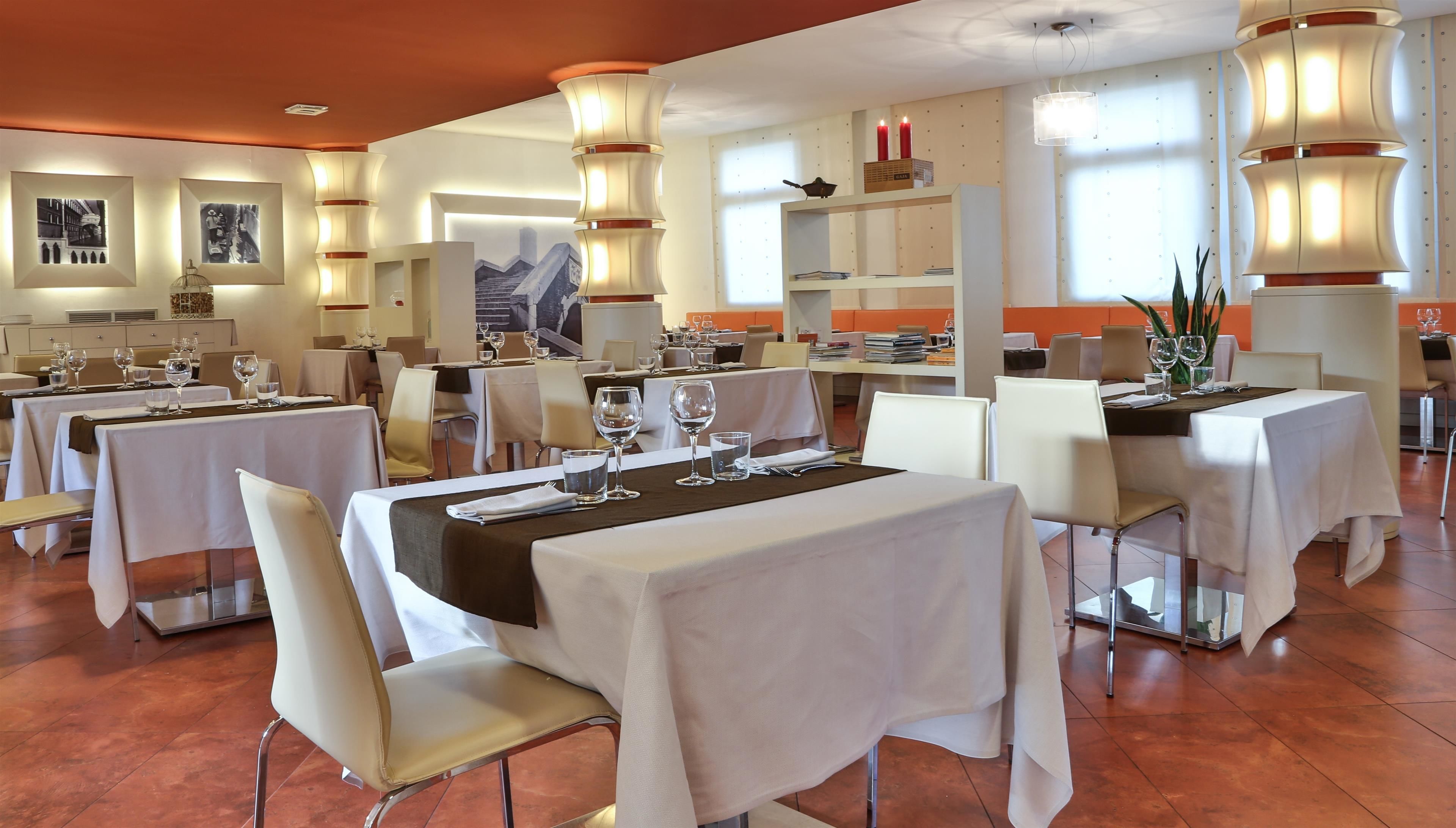 A restaurant with multiple tables set for dining, featuring a modern interior with white chairs and tables, and pendant lighting.