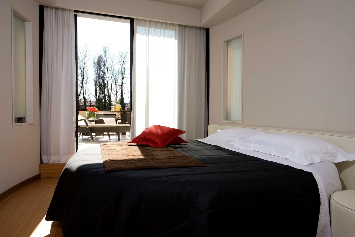 Une chambre d’hôtel avec un lit, un oreiller rouge, un banc en bois et une vue sur l’extérieur par une grande fenêtre.