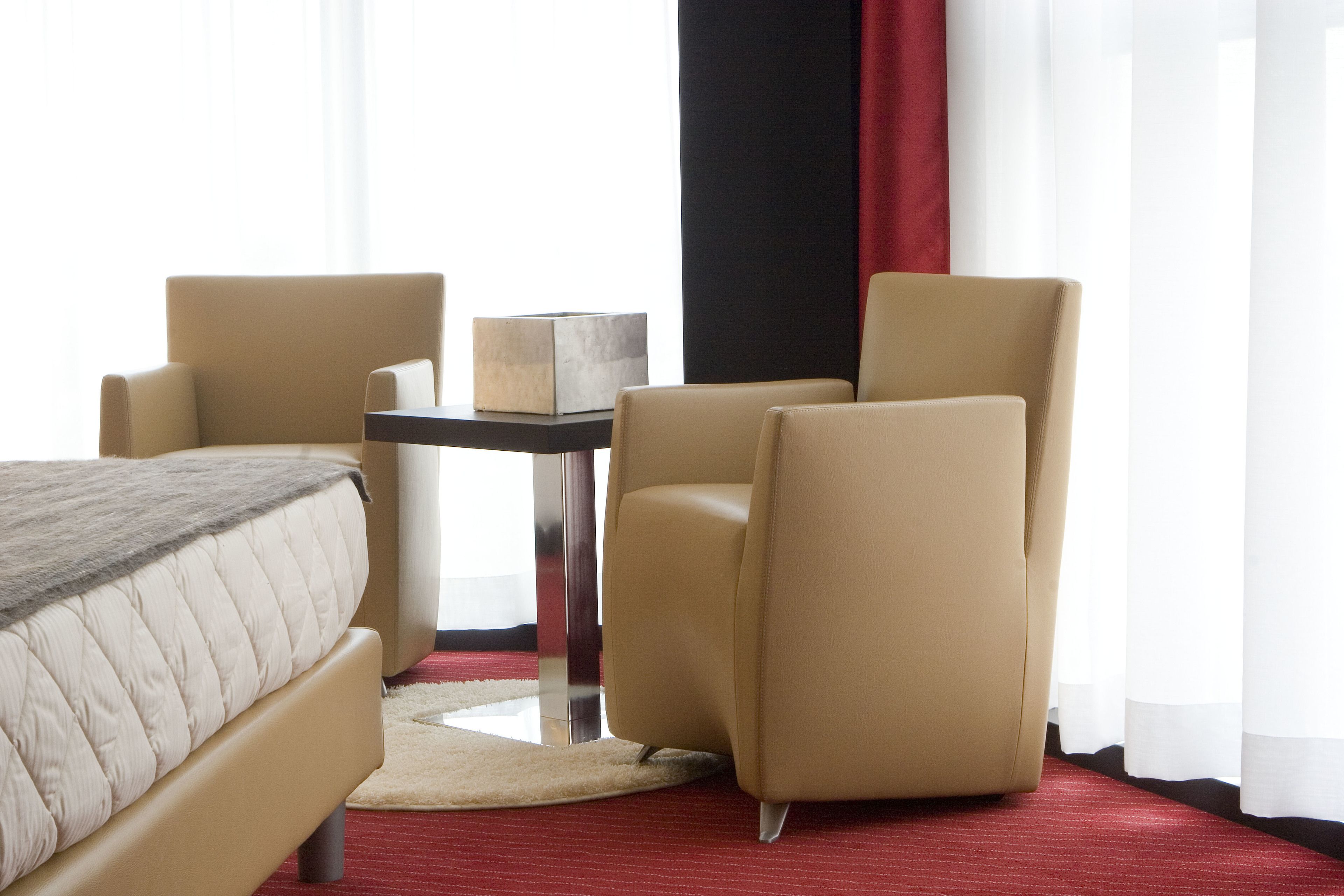 Une chambre d’hôtel avec un lit, deux fauteuils, une petite table et une moquette rouge.