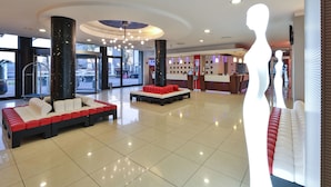 A spacious lobby with a modern design, featuring a central seating area with red and white cushions, a reception desk, and a large window offering a view of the outside.