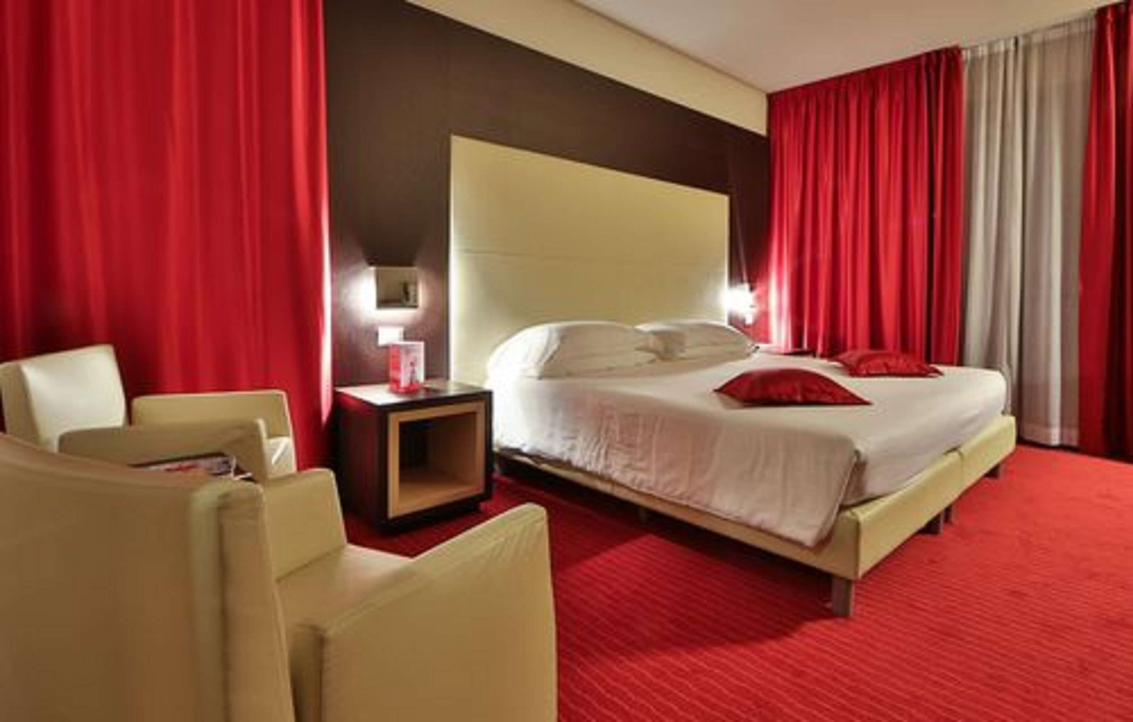 Habitación de hotel con una cama grande, una mesita de noche, una silla y cortinas rojas.