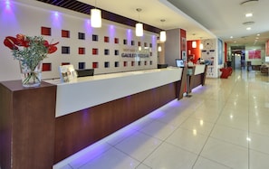 A hotel reception desk with a modern design, featuring a nameplate, a computer, and a vase with red flowers.
