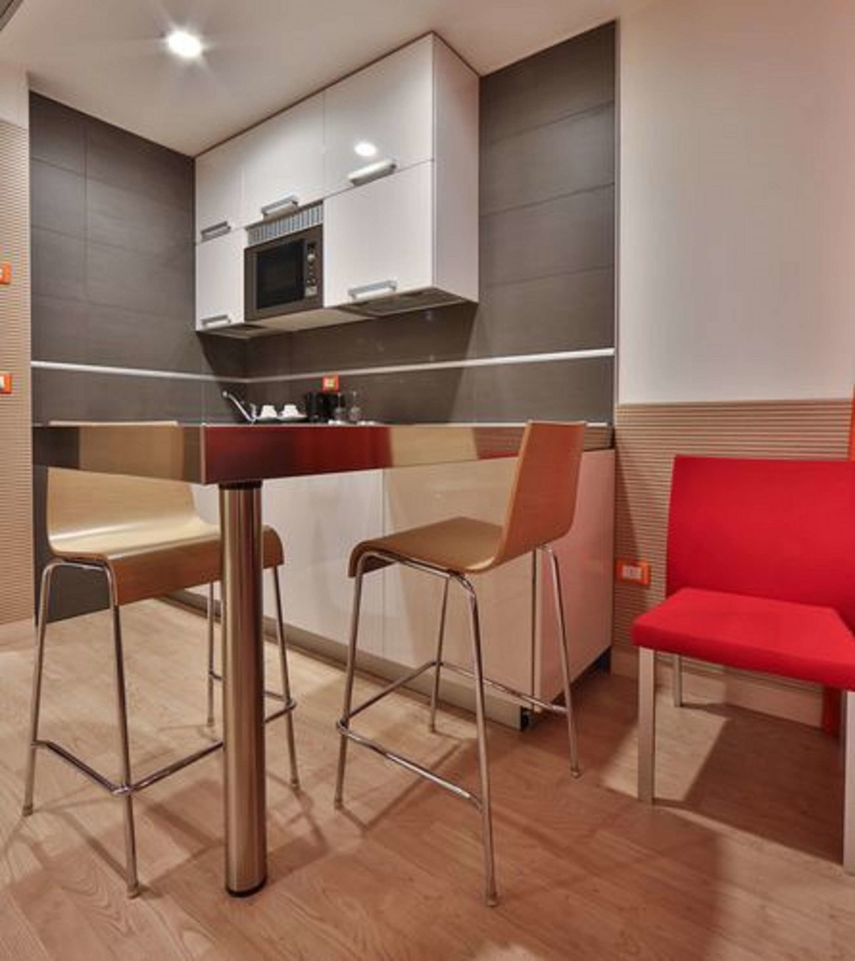 Una cocina moderna con microondas, un lavamanos y una silla roja. Hay dos taburetes de bar y una mesa de madera.