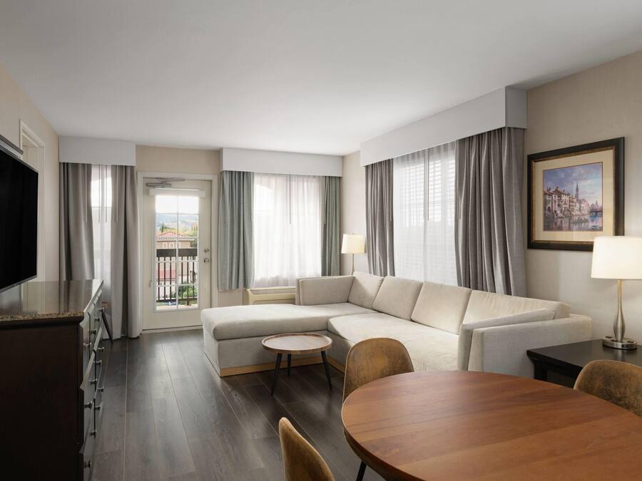 Quarto de hotel moderno com sofá, mesa de jantar, televisão e vista para o exterior.