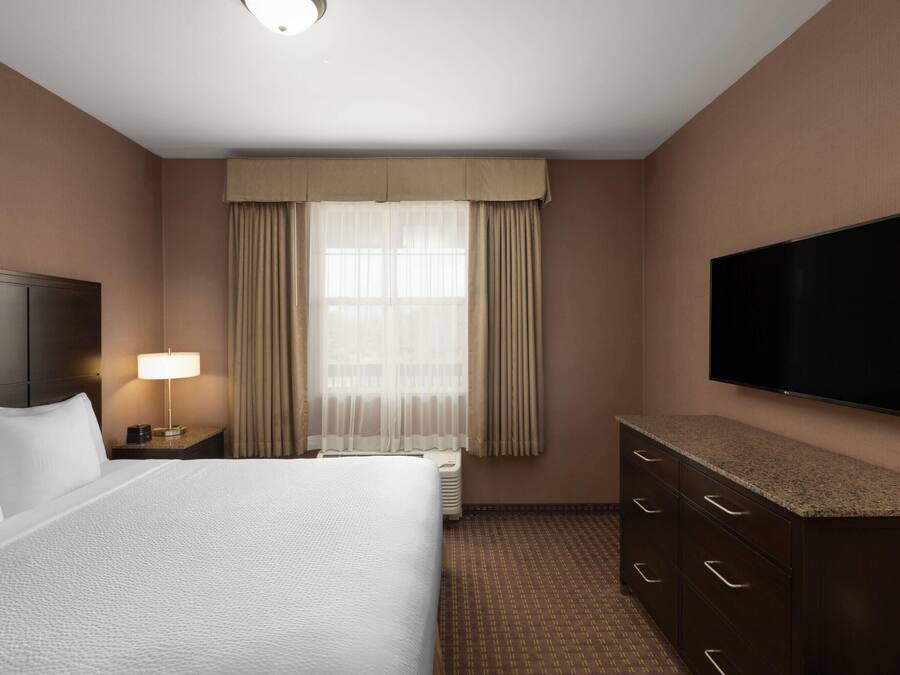 A hotel room with a large bed, a flat-screen TV, a nightstand with a lamp, a dresser, and a window with curtains.