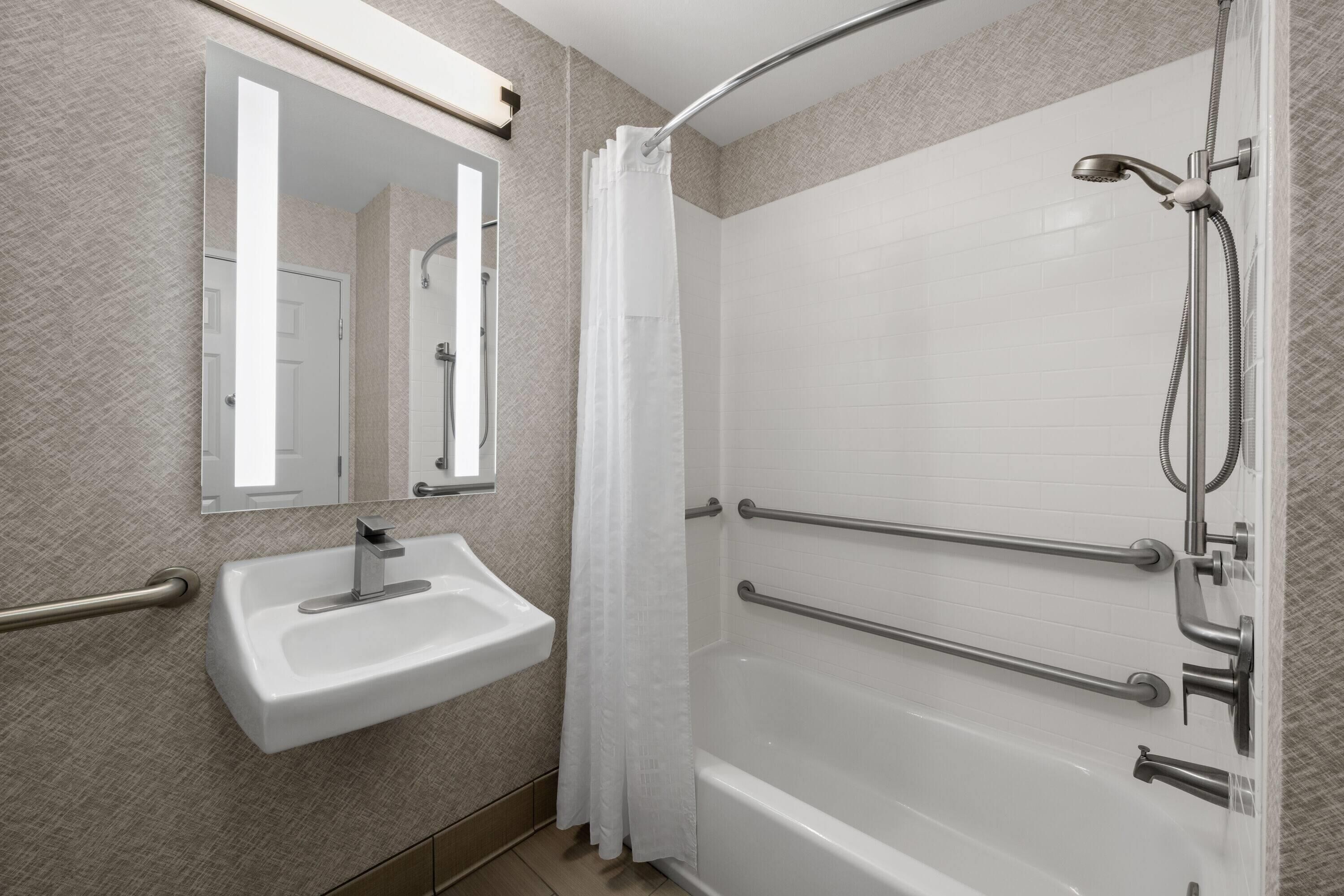 Un baño con lavamanos blanco, un espejo, ducha con puerta de vidrio y barras de apoyo.