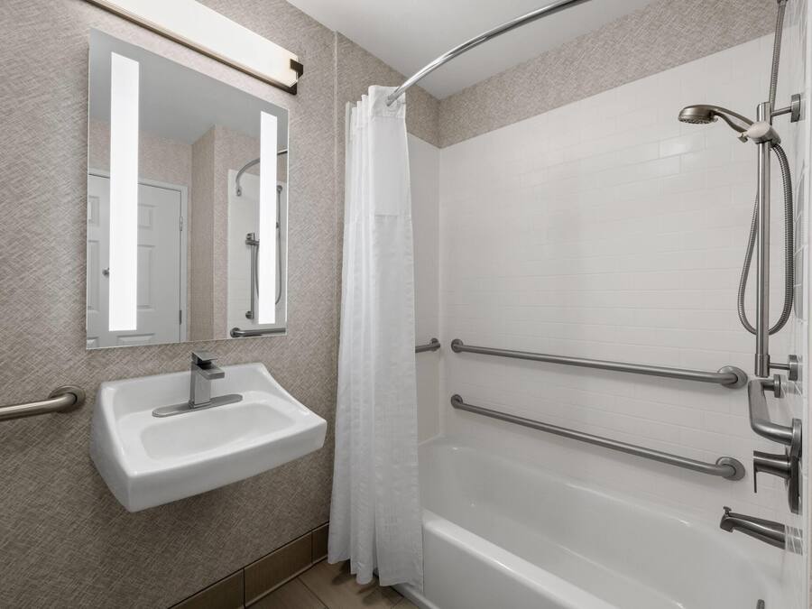 Un baño con lavamanos blanco, un espejo, ducha con puerta de vidrio y barras de apoyo.