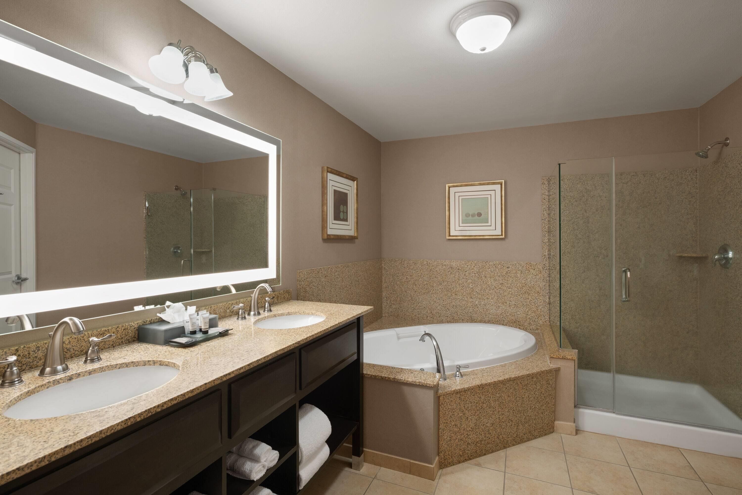 A modern bathroom with two sinks, a large bathtub, a glass shower enclosure, and a large mirror with integrated lighting.