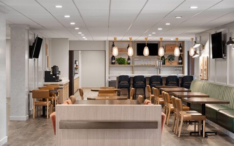 A modern restaurant with wooden tables and chairs, a central bar area, and a seating section with a green cushioned bench.