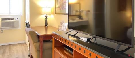 A hotel room with a wooden desk, a flat-screen TV, a lamp, a chair, and a dresser.