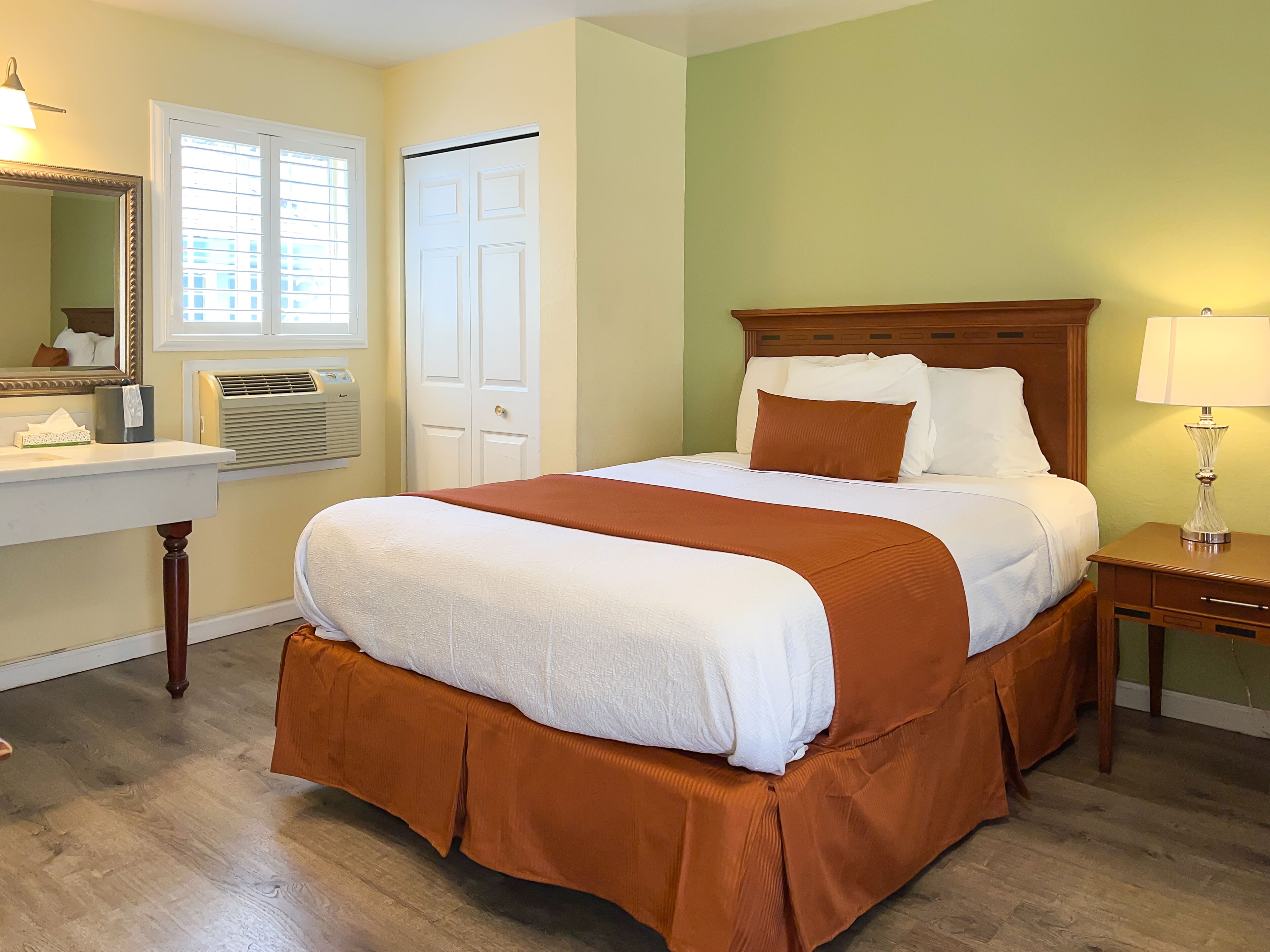 Une chambre d’hôtel bien aménagée, avec un grand lit, une table de chevet avec une lampe, un miroir et une fenêtre avec des stores.