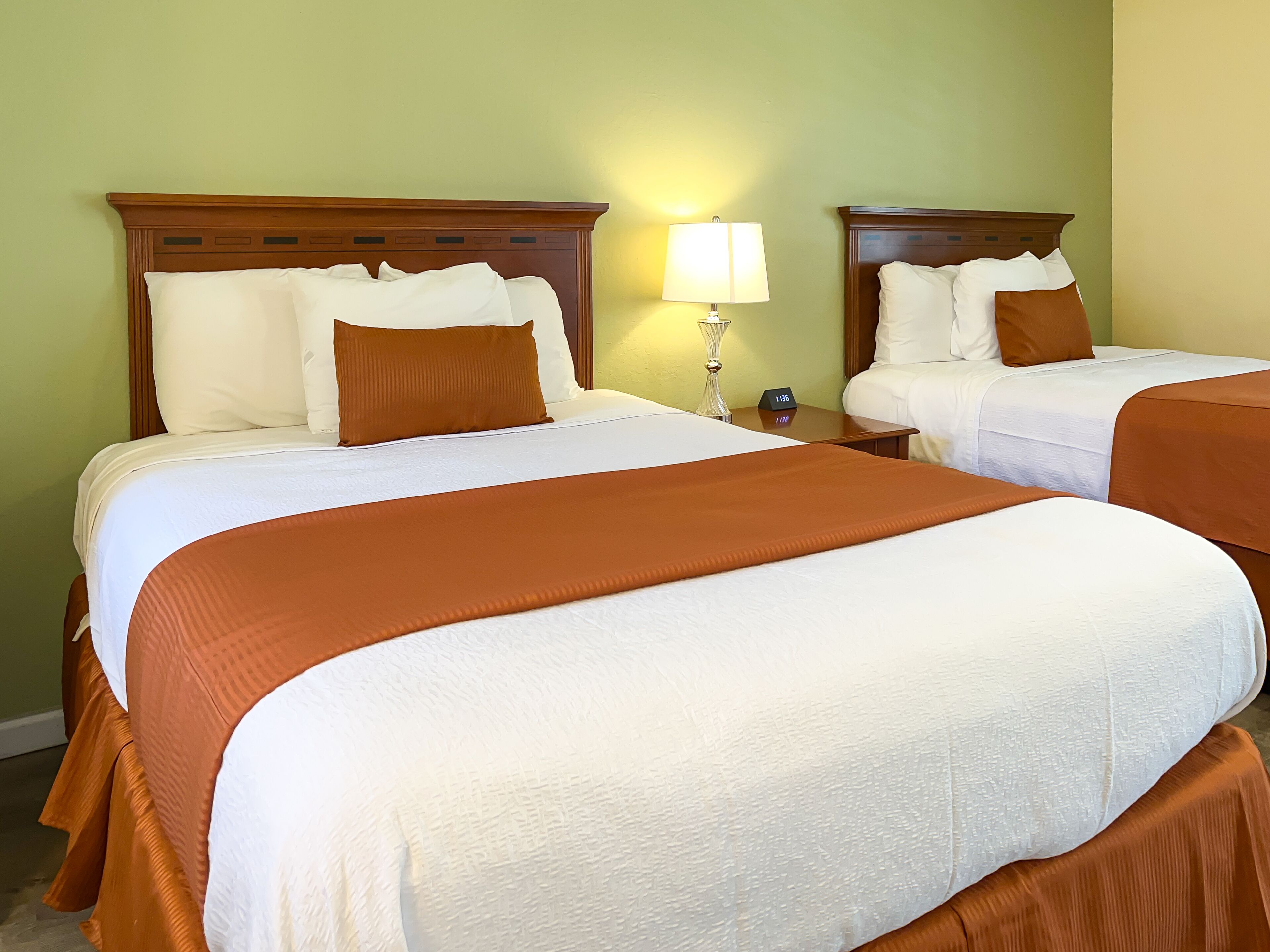 Habitación de hotel con dos camas, cada una con ropa de cama blanca y detalles en tonos marrones, una mesita de noche con lámpara y un reloj de pared.