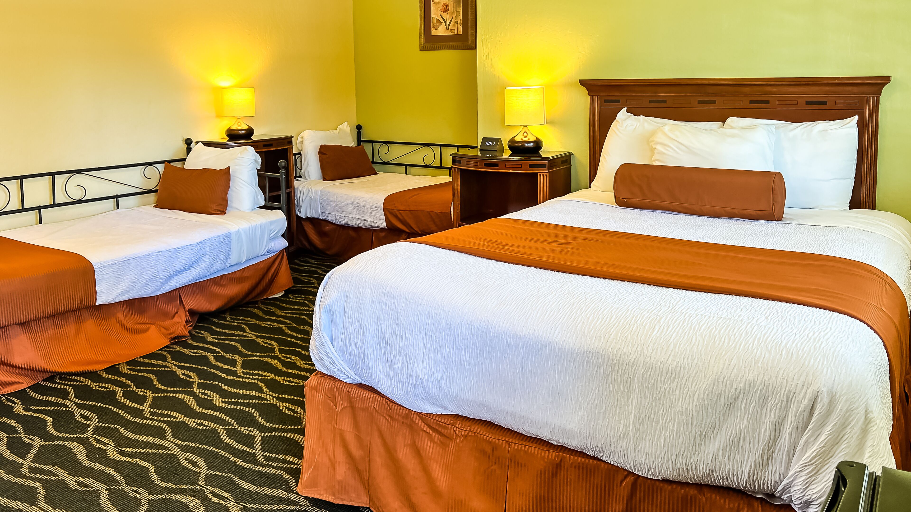 A hotel room with two single beds and one double bed, each with a wooden headboard and white bedding. There are bedside tables with lamps and a clock on the double bed. The room has a patterned carpet and green walls.