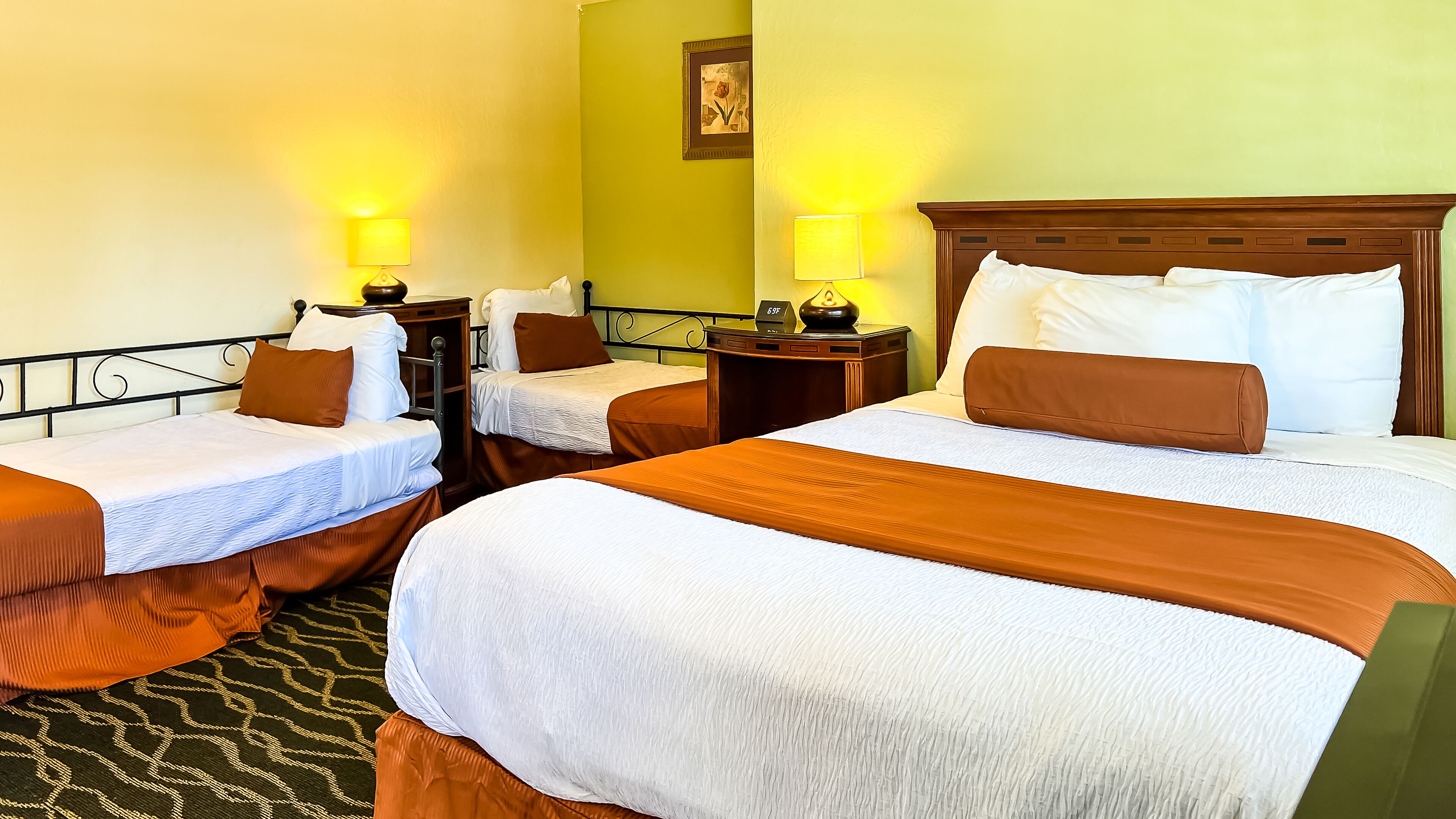 Habitación de hotel con dos camas individuales y una cama doble, cada una con cabecero de madera y ropa de cama blanca. Hay mesitas de noche con lámparas y un pequeño reloj sobre la cama doble. La habitación tiene una pared verde decorativa y un cuadro enmarcado sobre las camas individuales.