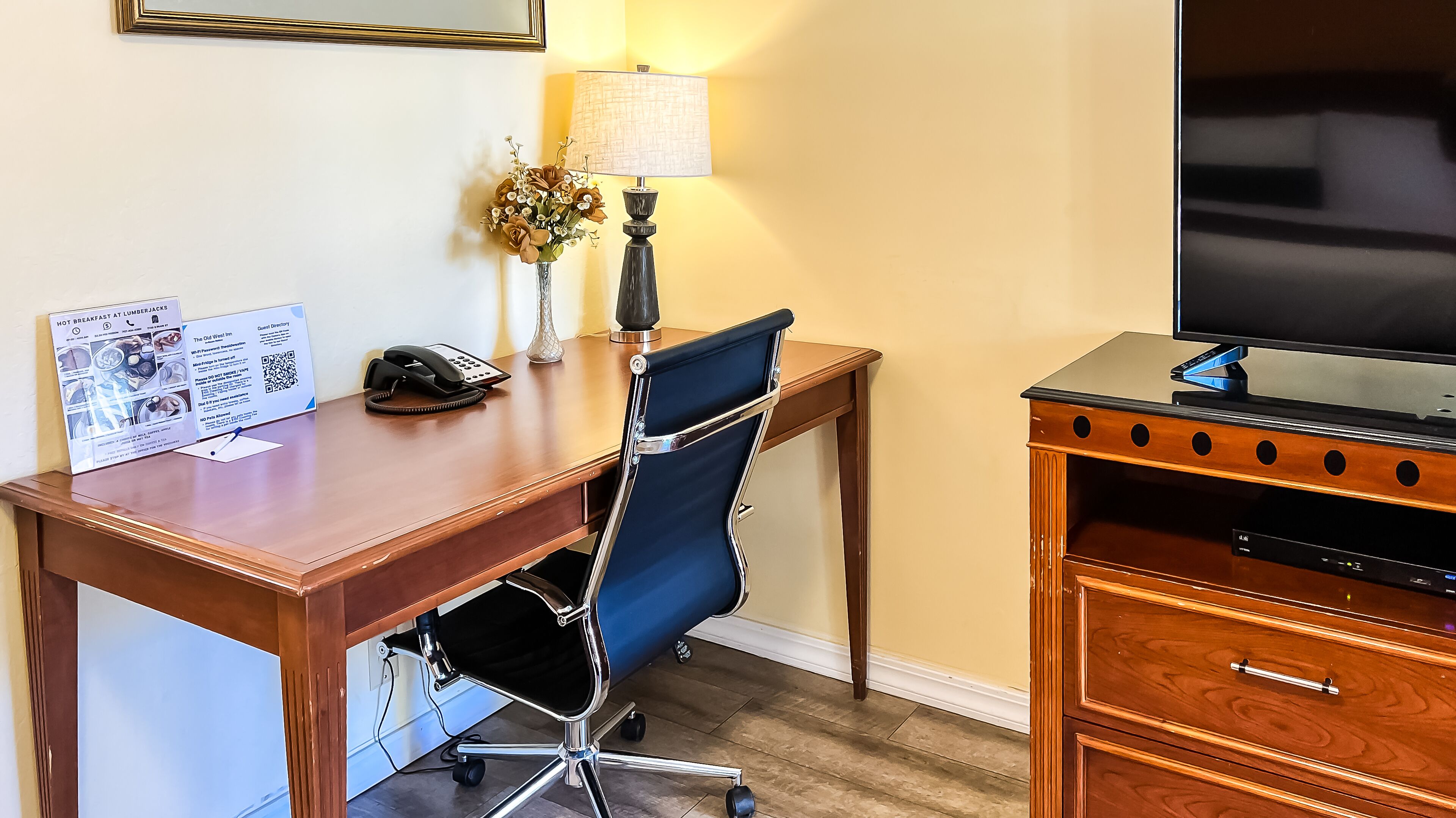 Een hotelkamer met een houten bureau, een blauwe bureaustoel, een lamp, een telefoon en een televisie op een houten standaard.