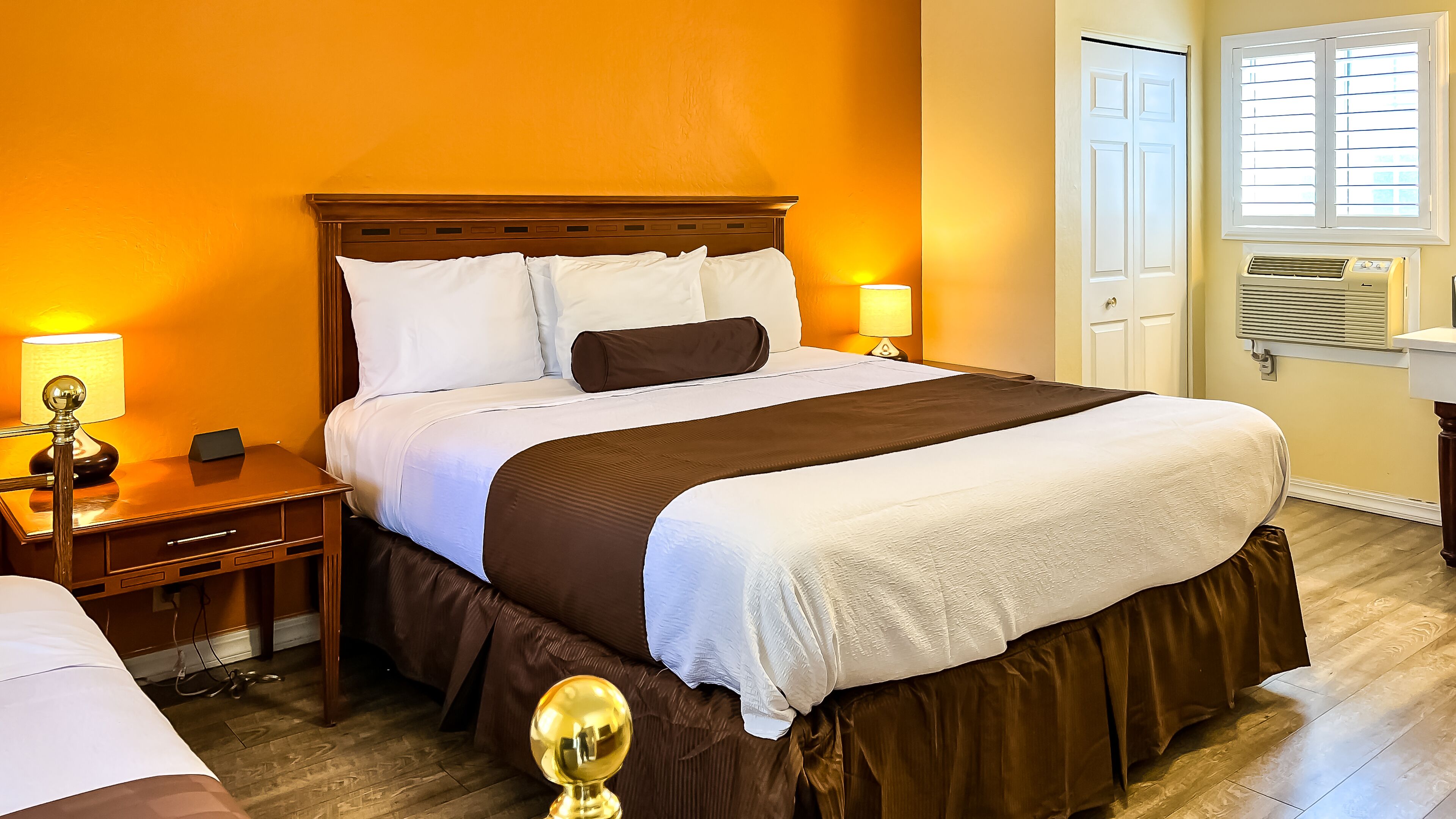 Une chambre d’hôtel avec un grand lit, une tête de lit en bois, deux tables de chevet avec des lampes, une fenêtre avec des stores et un appareil de climatisation.