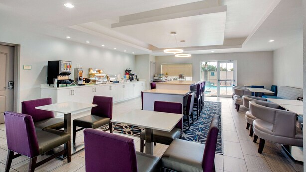 Un comedor moderno con mesas blancas y sillas moradas, una cocina al fondo y una alfombra estampada en el suelo.