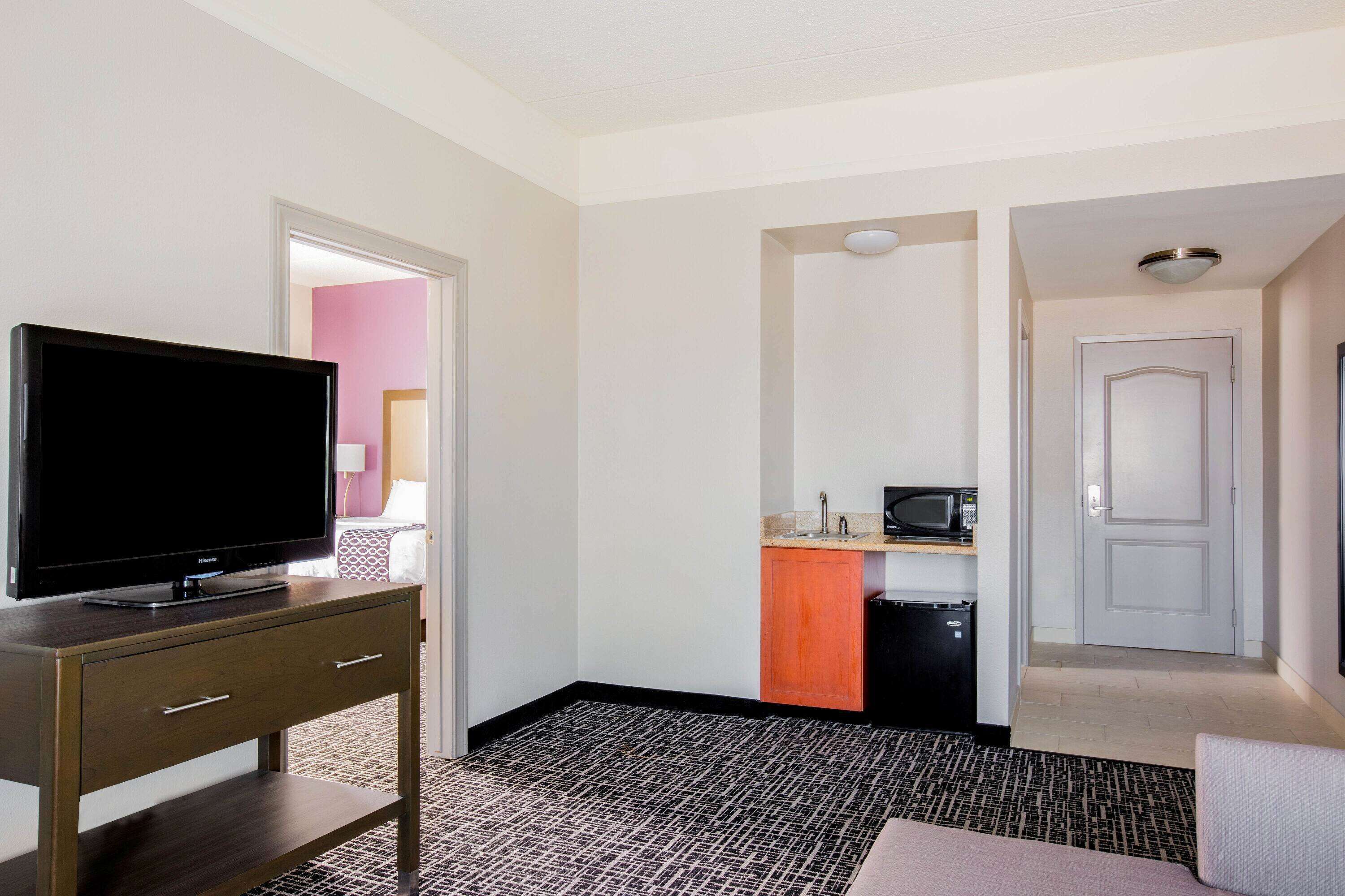 Habitación de hotel con televisión sobre un escritorio de madera, una kitchenette con microondas y una puerta que da a otra habitación.