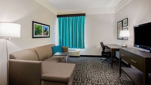 Una habitación de hotel con sofá, escritorio, televisor y ventana con cortinas.