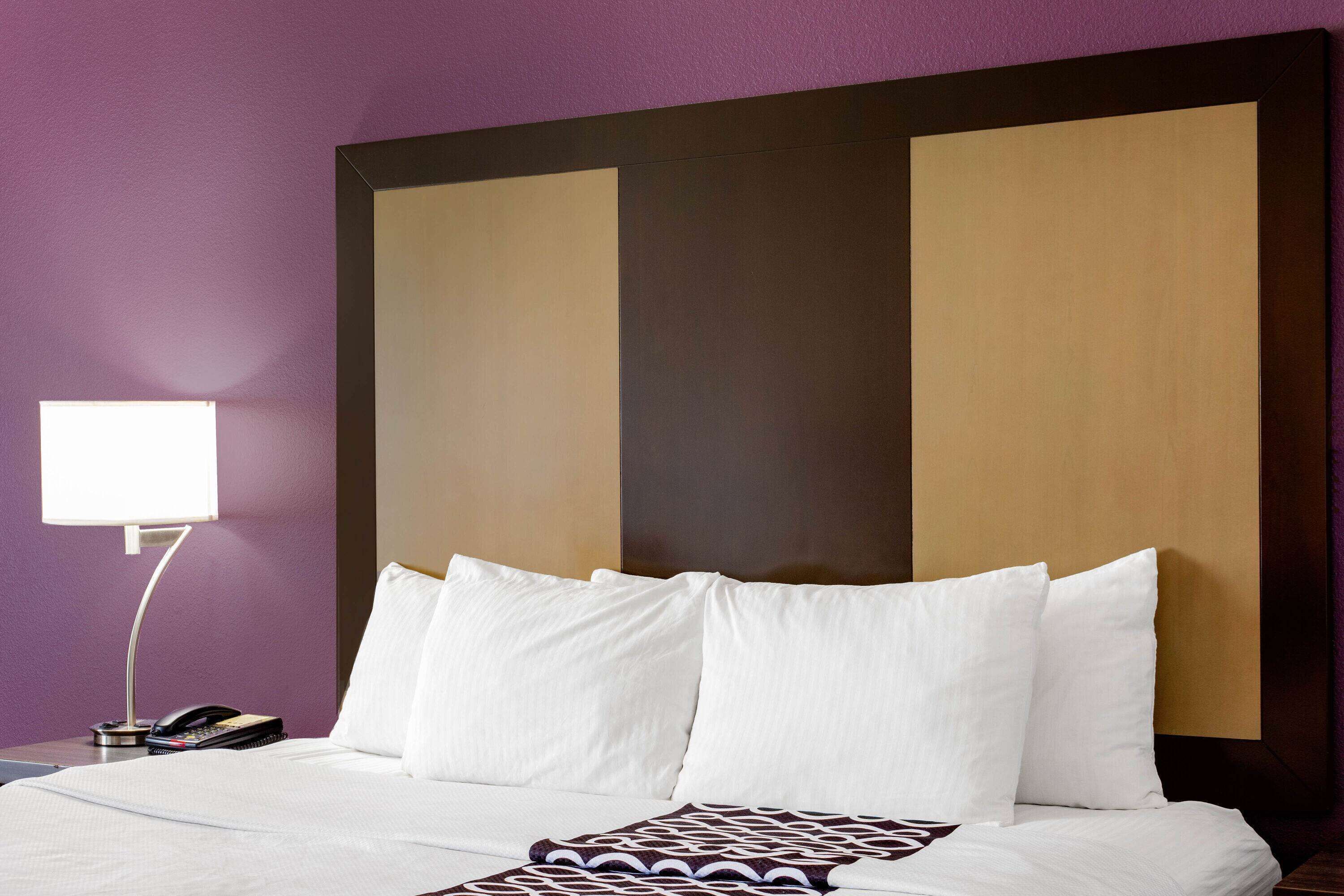 Habitación de hotel con una cama grande, ropa de cama blanca, cabecera con panel oscuro y lámpara de noche.