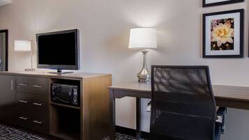 Habitación de hotel con televisor de pantalla plana, microondas, escritorio con lámpara, silla y dos cuadros en la pared.